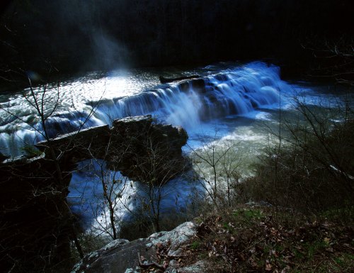 A serene waterfall cascades over rocks, surrounded by trees and mist, creating a tranquil natural scene.