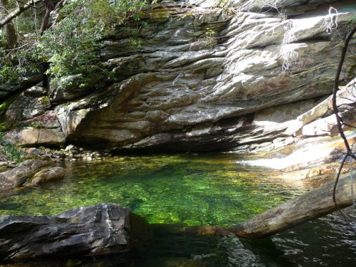 A serene natural pool surrounded by rocky cliffs and lush greenery, with sunlight reflecting on the water's surface.