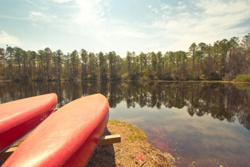 Two red kayaks rest on the shore by a calm lake, surrounded by trees and reflecting the blue sky.