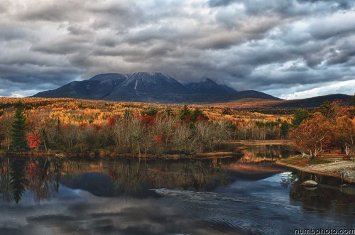 A serene lake reflects autumn foliage and a mountain under a dramatic, cloudy sky.