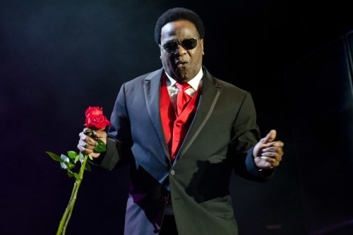 A performer in a black suit and sunglasses holds a red rose, singing passionately on stage.