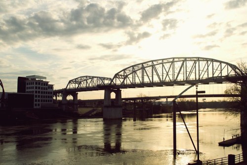 A bridge spans a river at sunset, with clouds reflecting on the water and buildings in the background.