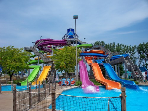 Colorful water slides at a water park, surrounded by trees and a clear blue sky, with a pool at the base.