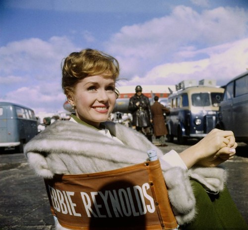 A smiling woman in a fur coat sits in a director's chair labeled "Debbie Reynolds," with vintage vehicles in the background.