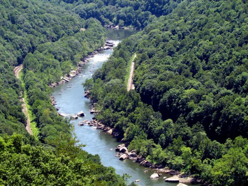 A winding river flows through lush green hills, surrounded by trees and rocky outcrops under a clear blue sky.