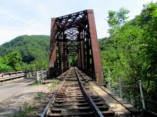 A rusted metal bridge spans a green valley, with train tracks leading into the distance under a clear blue sky.