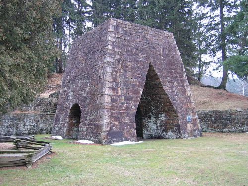 A stone structure with a triangular shape, surrounded by trees and grass, resembling a historic furnace or kiln.
