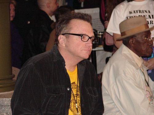 A man with glasses and a black jacket sits thoughtfully, next to another man in a hat, in a crowded setting.