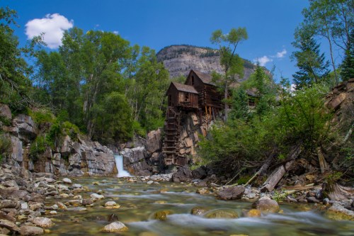 A rustic wooden mill beside a flowing river, surrounded by lush greenery and rocky cliffs under a clear blue sky.