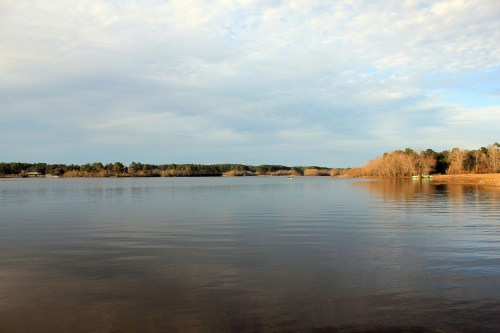 A calm lake reflecting the sky, surrounded by trees and a shoreline, under a partly cloudy sky.