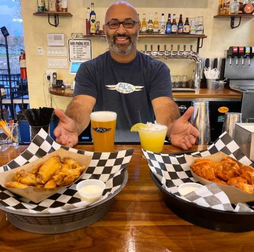 A smiling man stands behind a bar, showcasing two trays of chicken wings and drinks in a lively restaurant setting.