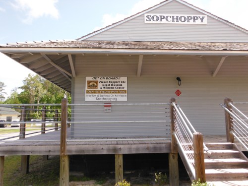 A wooden building with a sign reading "Sopchoppy" and a banner encouraging support for local events.