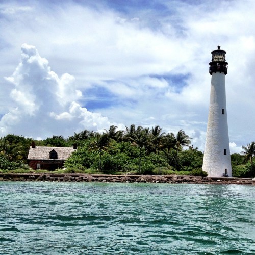 A tall white lighthouse stands beside a lush green landscape and a small building, under a partly cloudy sky.