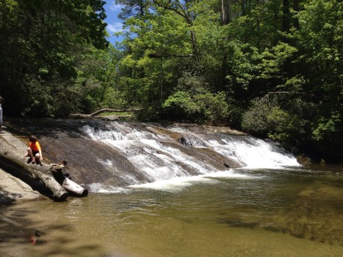 A serene waterfall cascades over rocks, surrounded by lush greenery and a clear pool, with people enjoying the scene nearby.