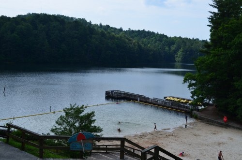 A serene lake surrounded by trees, with a sandy beach and a dock, where people are enjoying the water.