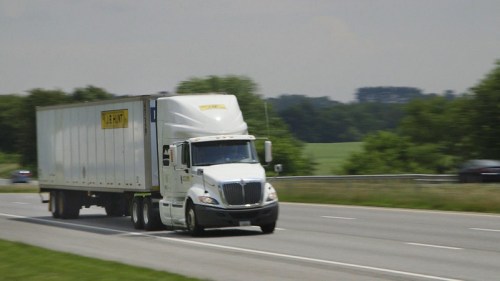 A white semi-truck with a yellow trailer drives on a highway, surrounded by green fields and trees.