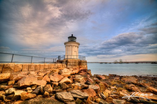 A lighthouse stands on rocky shore under a cloudy sky, with calm waters and distant structures in the background.