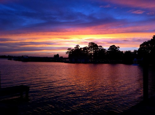A serene lake at sunset, with vibrant purple and orange skies reflecting on the water and silhouettes of trees.
