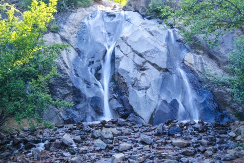 A serene waterfall cascading down rocky cliffs, surrounded by lush greenery and scattered stones at the base.