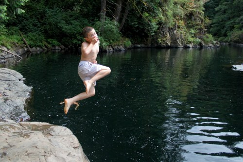 A boy jumps joyfully into a calm, dark pool surrounded by lush greenery and rocky edges.