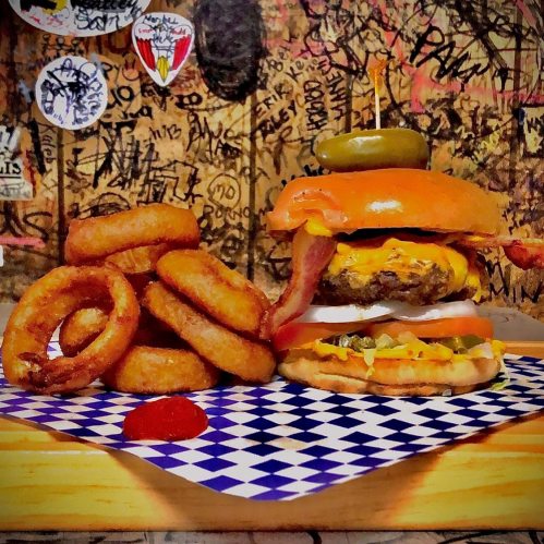 A stacked cheeseburger with bacon, pickles, and onion rings on the side, served on a checkered paper.