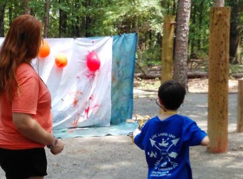 A woman and a child stand outdoors, preparing to pop balloons on a target with a slingshot. Trees surround them.