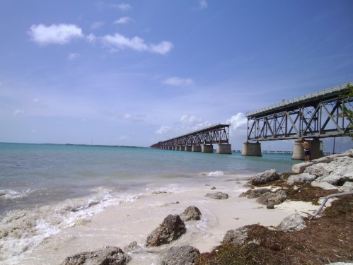 A scenic view of a bridge extending over calm turquoise waters, with sandy shores and rocky outcrops in the foreground.