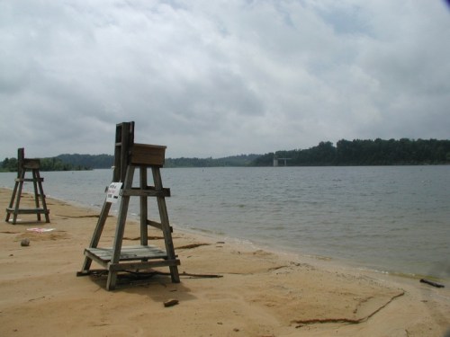 Two wooden lifeguard stands on a sandy beach by a calm lake under a cloudy sky.