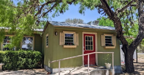 A small green building with a red door and an "open" sign, surrounded by trees and shrubs.
