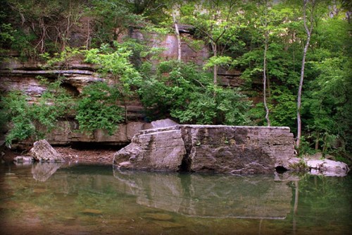 A serene landscape featuring a rocky outcrop beside a calm, reflective body of water, surrounded by lush greenery.