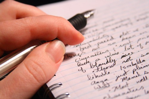 A close-up of a hand holding a pen, writing notes in a notebook filled with handwritten text.