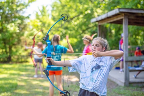 A girl aims a blue bow while practicing archery outdoors, with other participants in the background.
