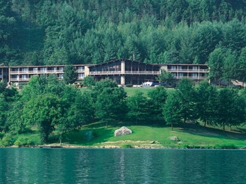 A lakeside hotel surrounded by lush greenery and trees, with a clear blue water view in the foreground.