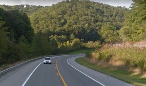 A winding road curves through lush green hills under a clear sky, with a car driving along the highway.