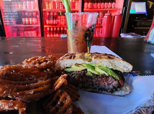 A juicy burger with avocado on a bun, served with crispy waffle fries and a drink in a vibrant bar setting.