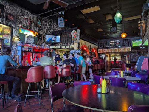 A lively bar interior with colorful seating, patrons at the counter, and vintage decor on the walls.