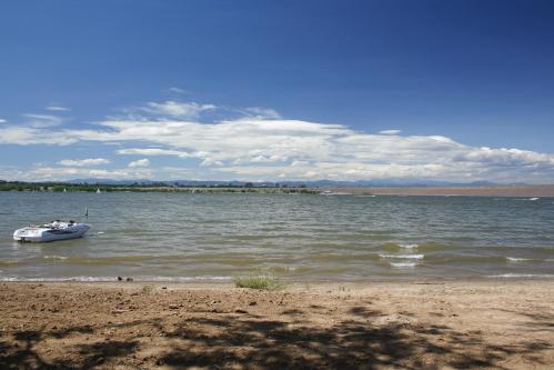 A serene lakeside view with gentle waves, a sandy shore, and distant mountains under a blue sky with fluffy clouds.