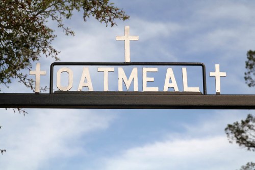 Sign reading "OATMEAL" with decorative crosses above it, set against a blue sky and tree branches.