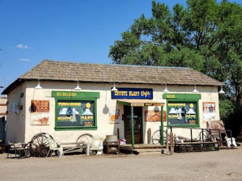 A quaint burger cafe with colorful signs, surrounded by rustic decor and greenery under a clear blue sky.