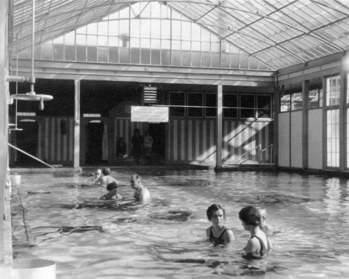 Black and white photo of a large indoor pool with people swimming and relaxing, surrounded by glass walls.