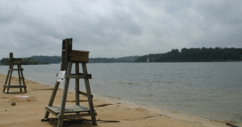 Two wooden lifeguard stands on a sandy beach by a calm lake, with overcast skies and trees in the background.