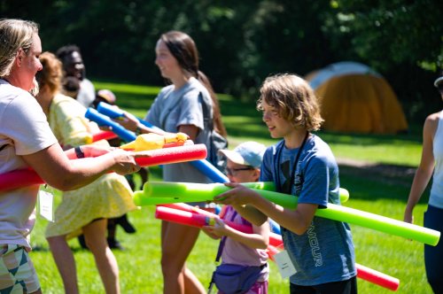 Children and adults play with colorful pool noodles in a sunny outdoor setting, with tents in the background.