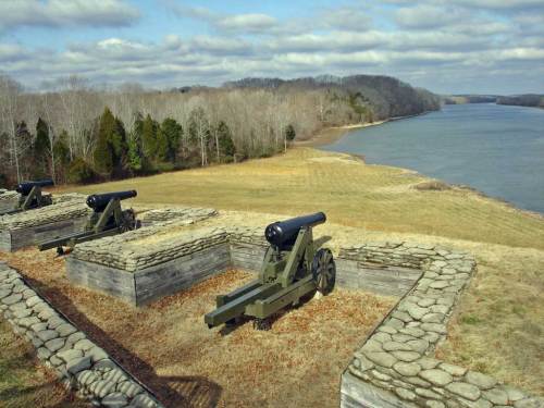 Historic cannons positioned on a grassy overlook by a river, surrounded by trees and under a cloudy sky.