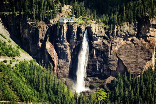 A stunning waterfall cascades down a rocky cliff, surrounded by lush green trees and a house perched above.