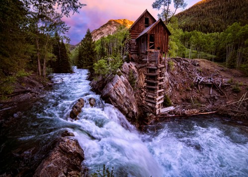 A rustic wooden cabin perched on a rocky outcrop beside a rushing river, surrounded by lush trees and mountains at sunset.