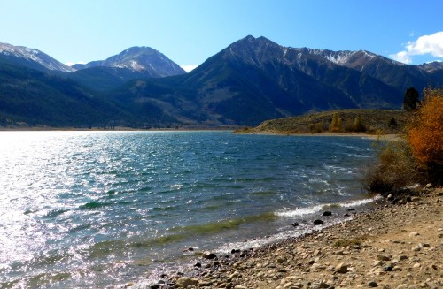 A serene lake surrounded by mountains, with clear blue water and a rocky shoreline under a bright sky.