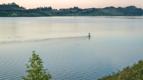 A person paddleboarding on a calm lake surrounded by green hills at sunset.