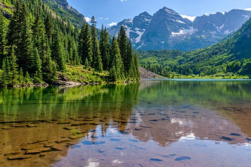 A serene lake surrounded by lush green trees and majestic mountains, reflecting the clear blue sky.