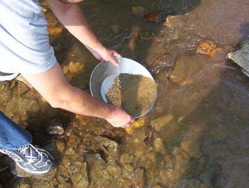 A person pans for minerals in a shallow stream, holding a metal pan filled with gravel and water.
