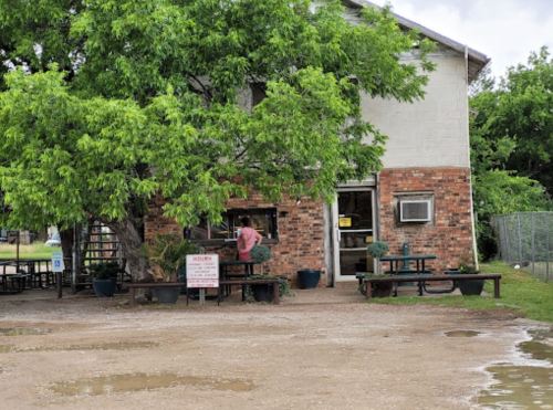 A small brick building with a large tree in front, featuring outdoor seating and a sign displaying business hours.